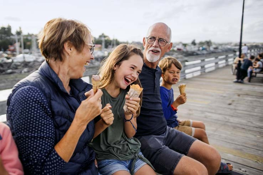 Großeltern mit Enkelin und Enkel beim Eis auf einer Bank an einem Hafen. Alle lachen ausgelassen.