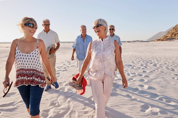 Eine Gruppe von Rentnerinnen und Rentnern genießt unbesorgt ihren Ruhestand bei einem Spaziergang am sonnigen Strand.